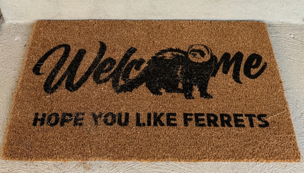 Brown welcome mat with an image of a ferret as part of the word Welcome, followed by the words, hope you like ferrets. 